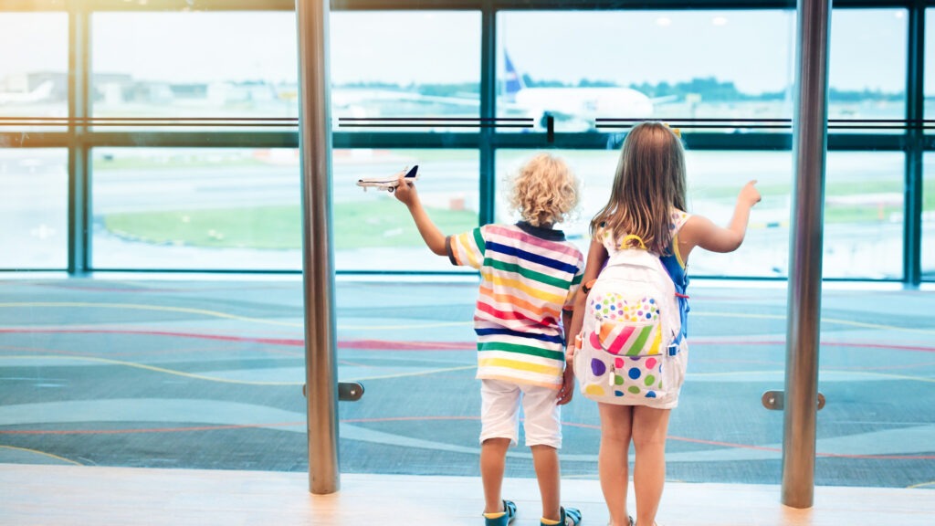 A boy and a girl looking at an airplane through the airport window. The boy is holding a mini airplane and the girl is wearing a colorful backpack. One of th must-haves when traveling with kids