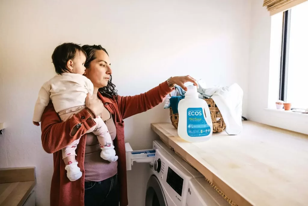 A woman in a laundry roomm holding a baby and loading the washing machine. She is using ECOS non toxic laundry detergent