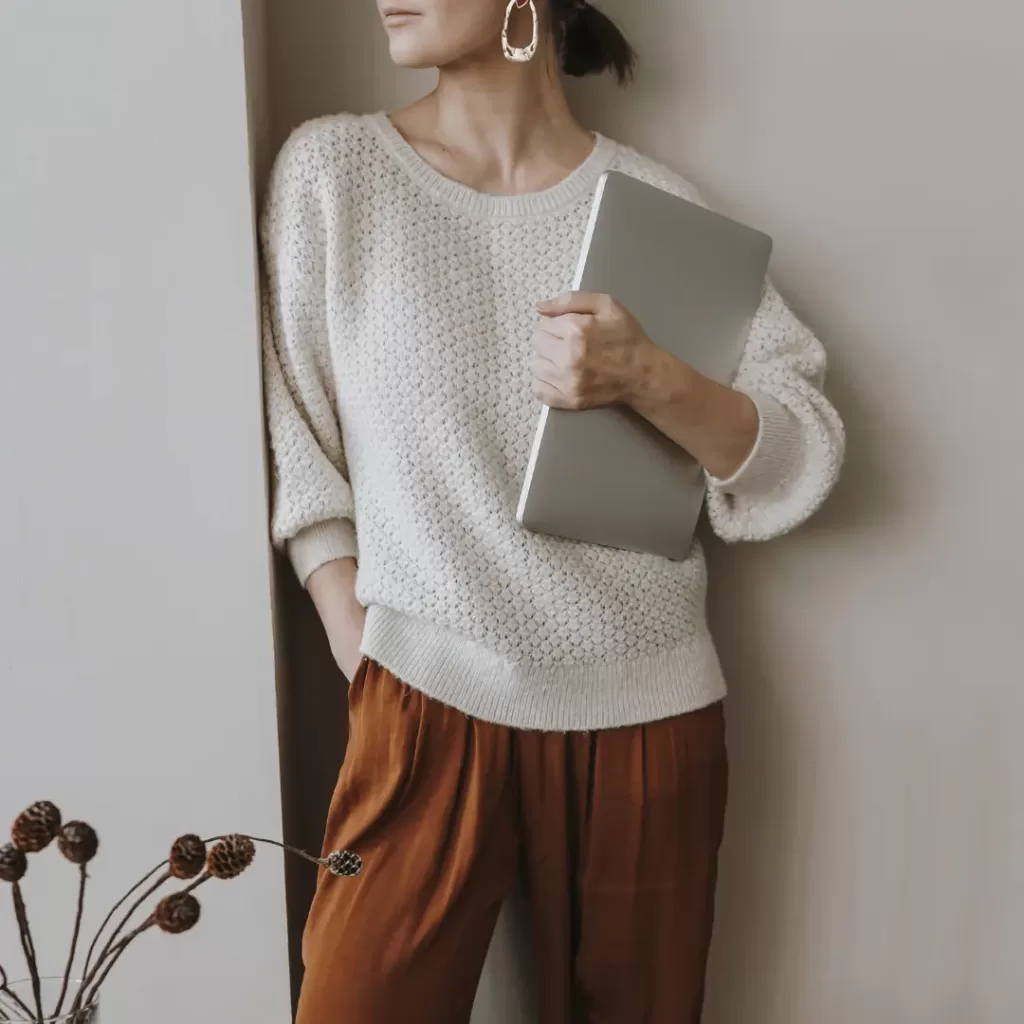 A woman in a white swater and rust pant. Holding her laptop. Time Management for working moms is also to help get more work done