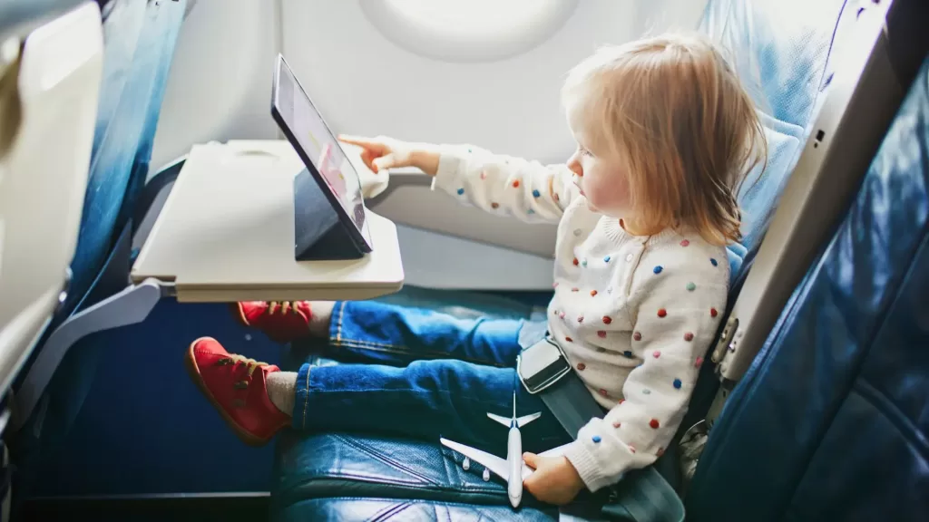 a toddler seating in a plane and playing on her tabler. Oe of th must-haves on a packing list for toddlers